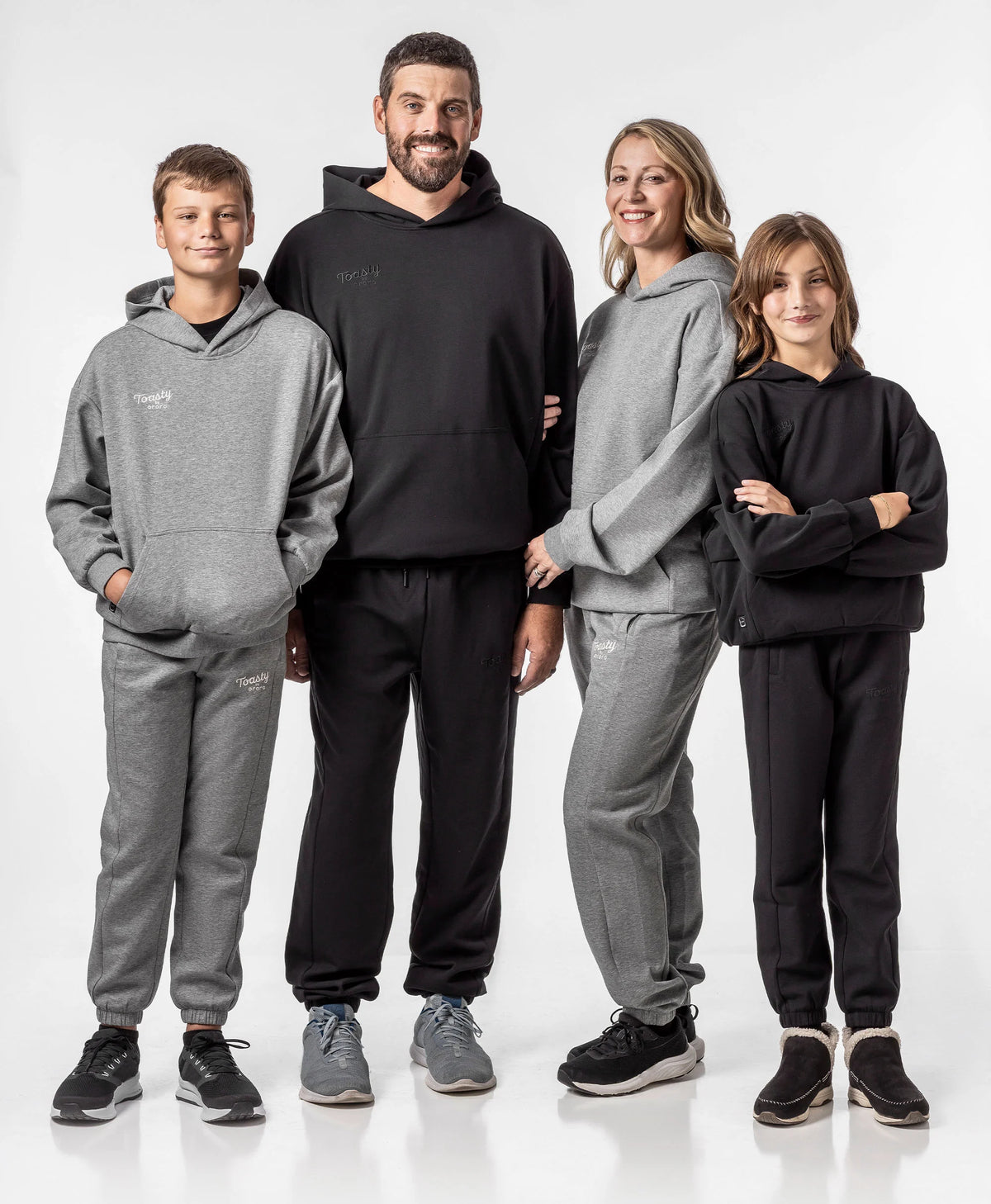 A four people family standing together in matching Toasty Unisex Heated Pullover Hoodies and sweatpants sets, with two adults and two children wearing a mix of black and flecking gray loungewear against a white background.
