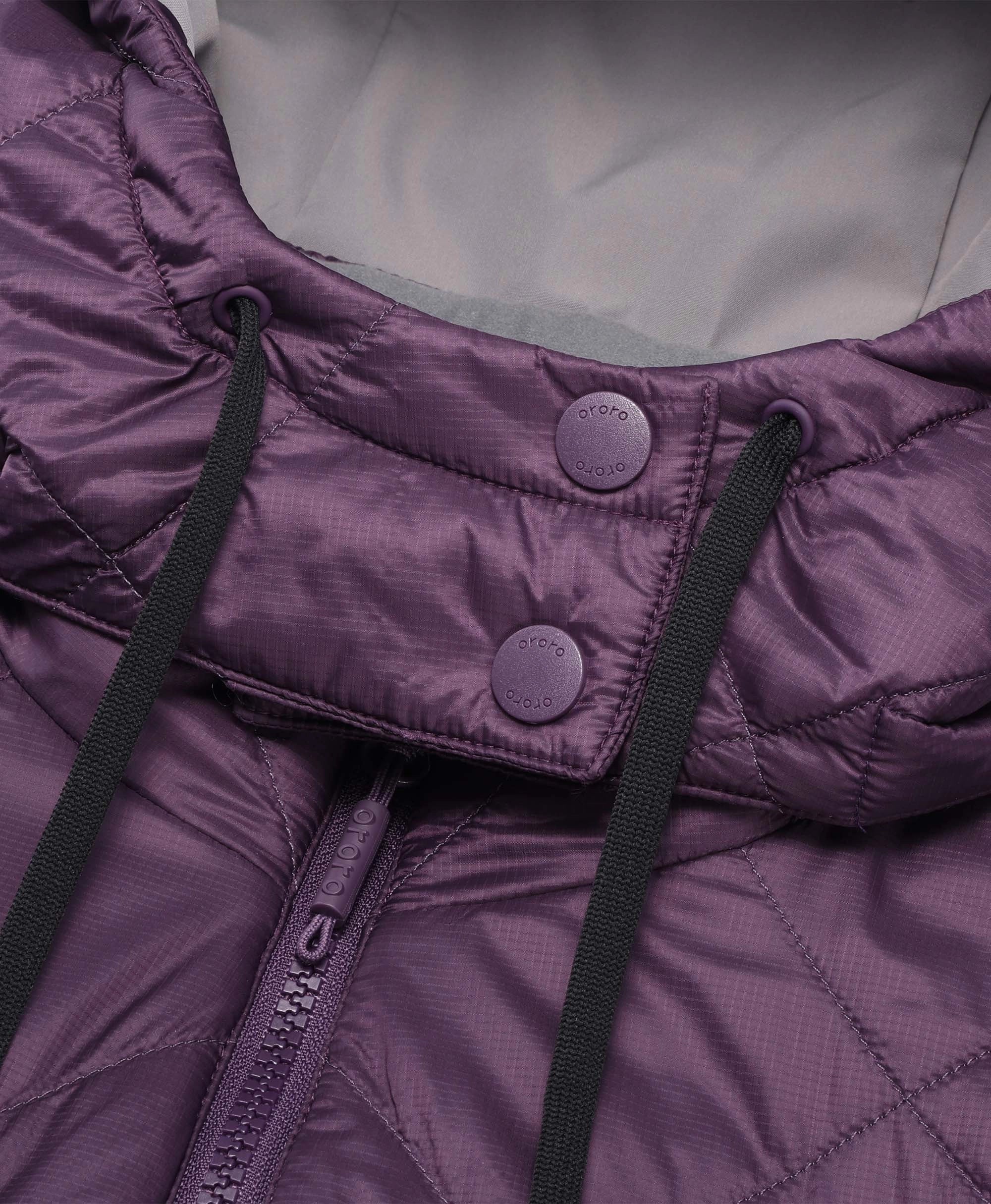 Close-up of a purple quilted puffer jacket showing the collar, snap-button flap, black drawstrings, and a zipper.