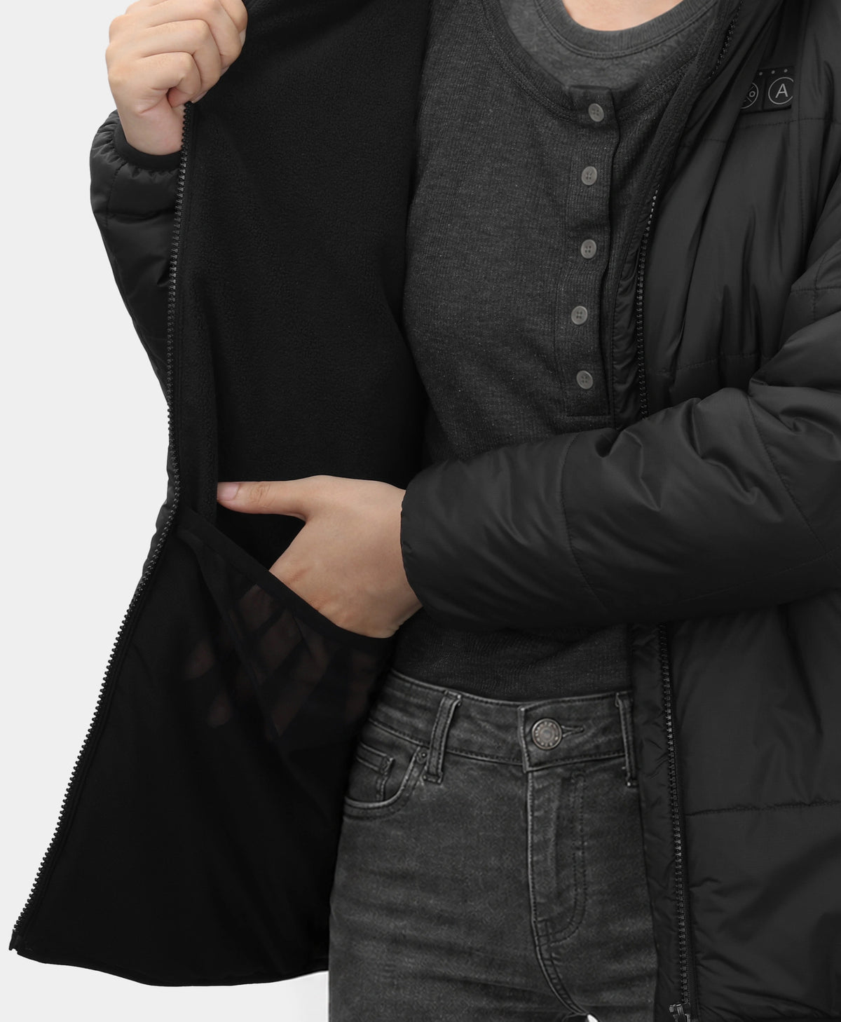 Close-up of a person wearing a black puffer jacket over a dark gray henley shirt and dark gray jeans, with one hand opening the jacket and the other hand reaching into a side pocket.