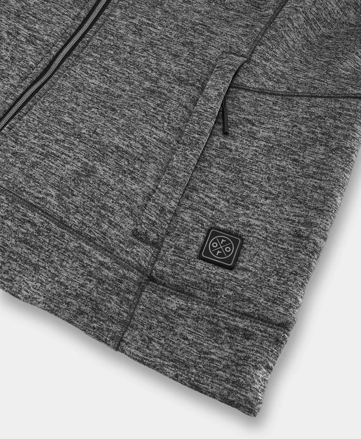 Close-up of a dark heather gray zippered garment with a small square black brand patch on the front pocket, photographed on a light gray background.