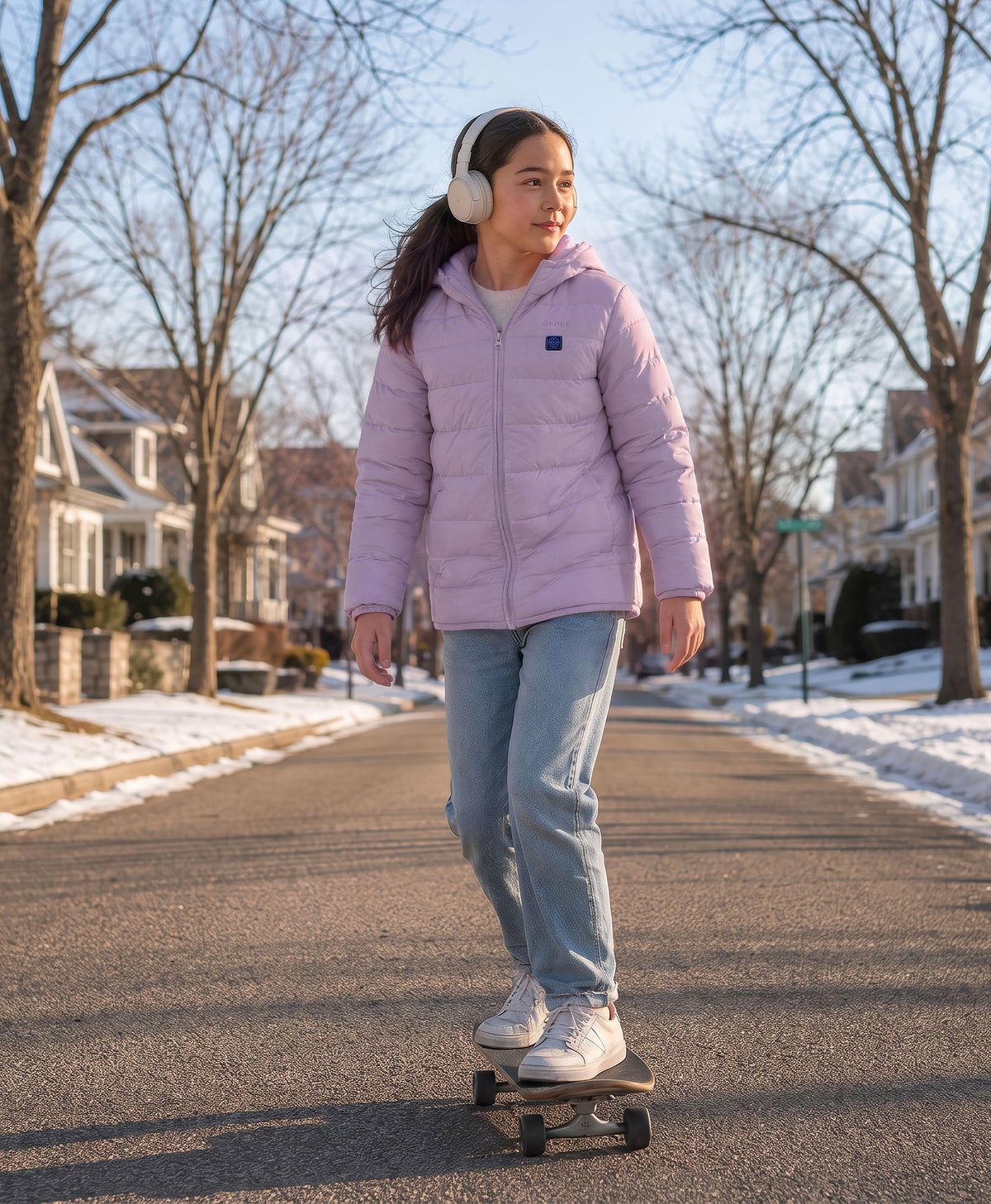 A young girl wearing ororo light purple puffer jacket, riding a skateboard down a snowy residential street.