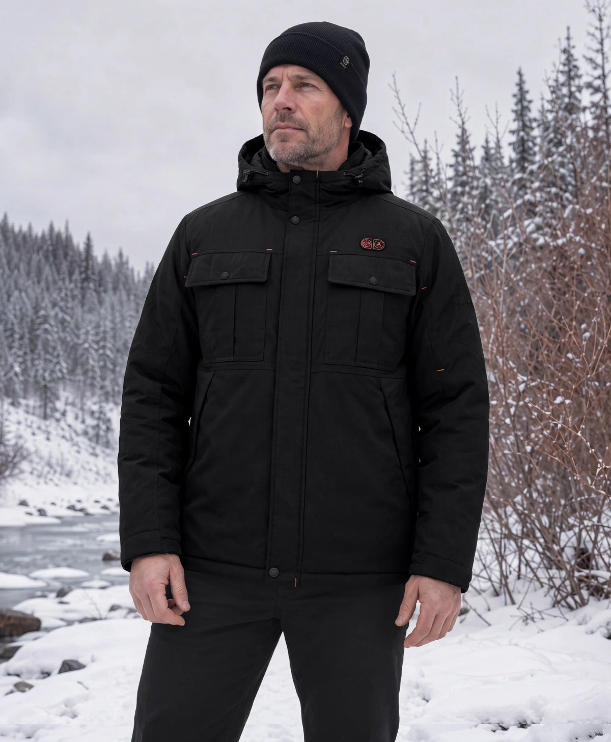 A man wearing a ororo SureWarm® jacket and black beanie standing outdoors in a snowy landscape.