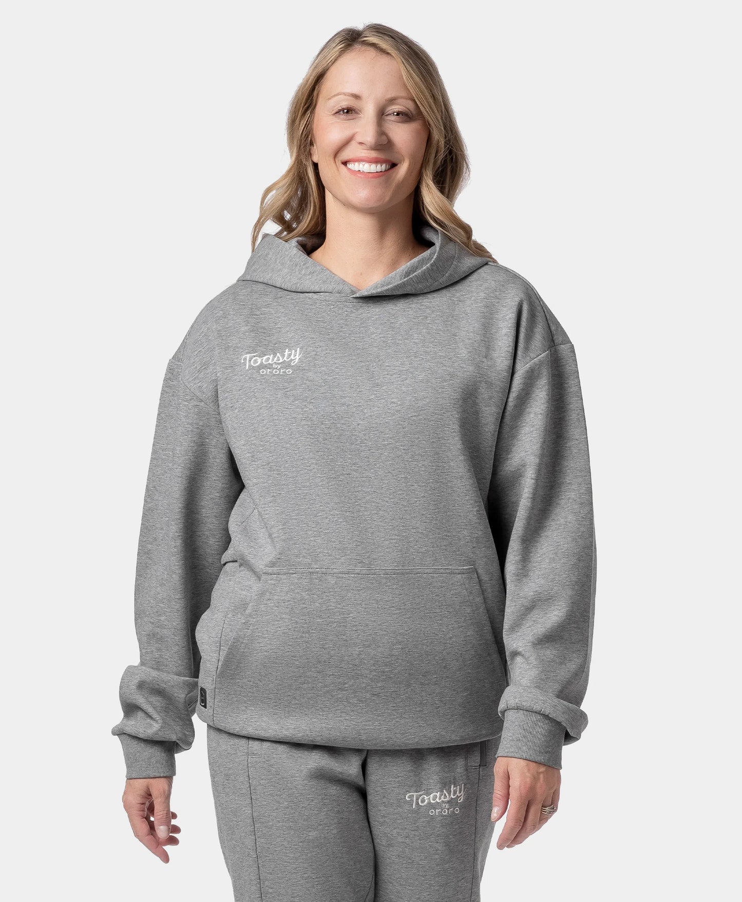 A smiling woman wearing a flecking gray Toasty Unisex Heated Pullover Hoodie and matching sweatpants with a small white "Toasty by ororo" logo printed on the left chest and upper thigh, photographed against a plain off-white studio background.