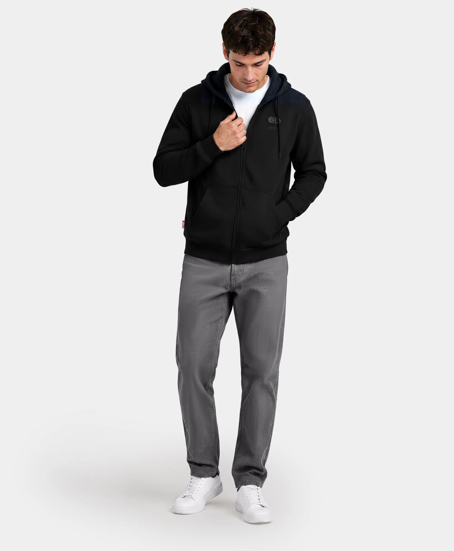 A man standing with hands in pockets wearing the black SureWarm® Unisex 6-Zone Fleece Heated Hoodie with gray pants