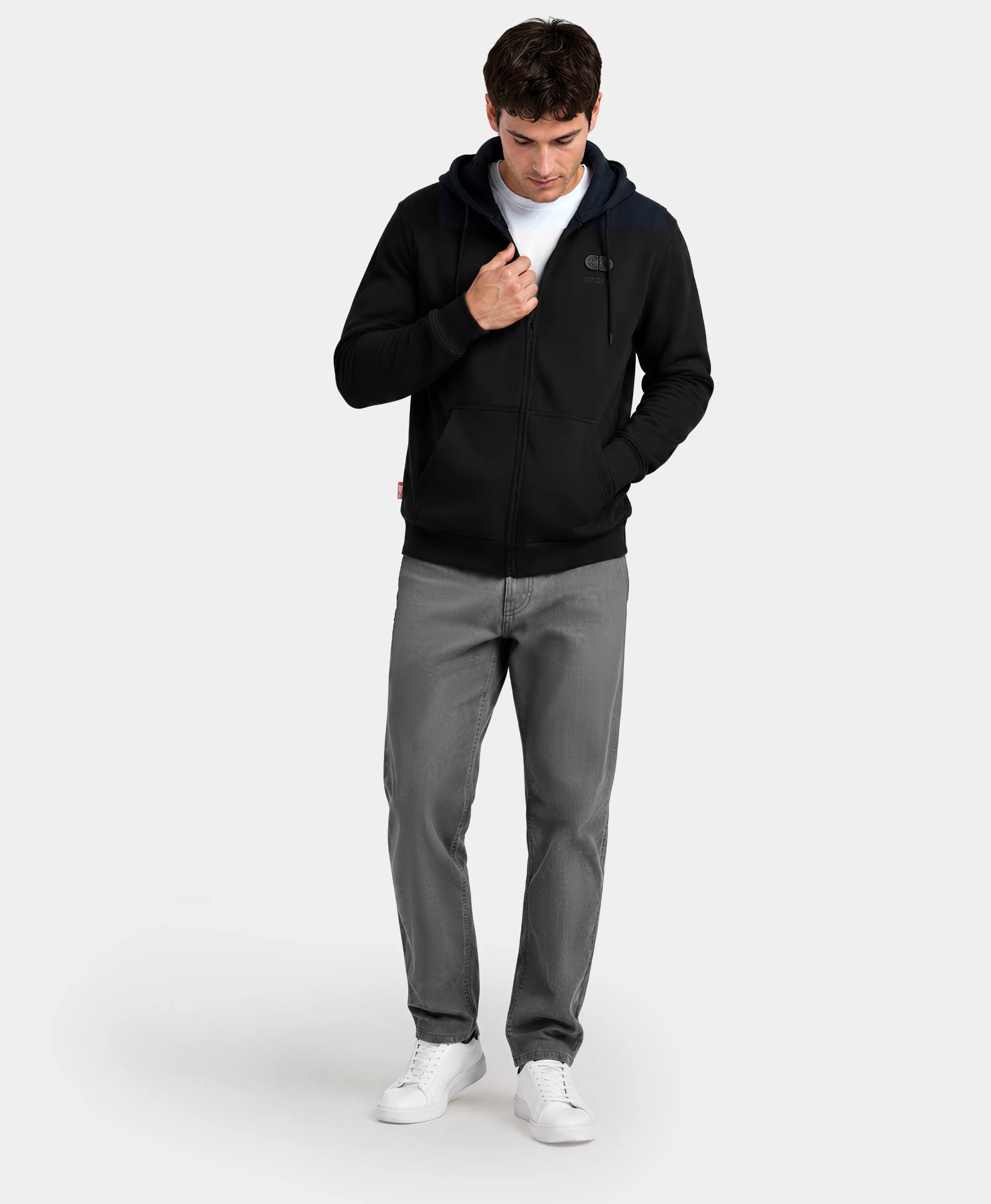 A man standing with hands in pockets wearing the black SureWarm® Unisex 6-Zone Fleece Heated Hoodie with gray pants