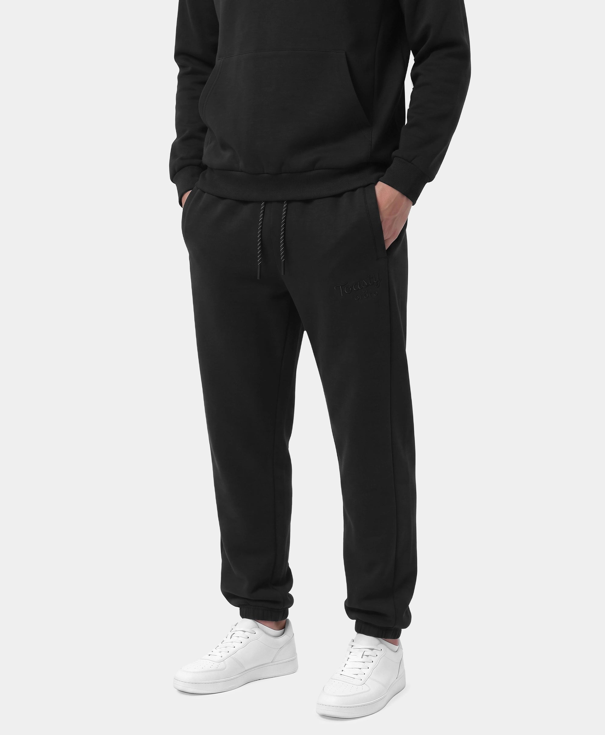 A person wearing black jogger sweatpants with a drawstring waist, paired with a black hoodie and white sneakers, shown against a light gray studio background.